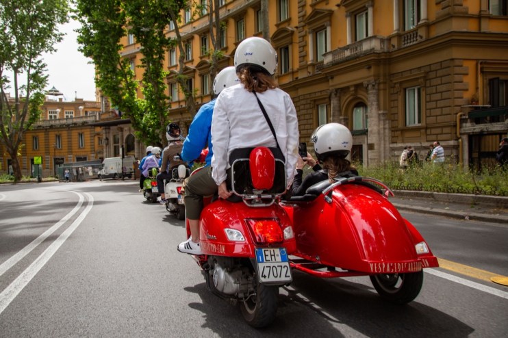 Tourists on a red Vespa with sidecar ride through Rome, Italy, past historic buildings.
