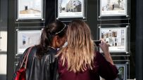 Two women view property listings in an estate agents window, one takes a photo with her phone. Real estate, stamp duty.
