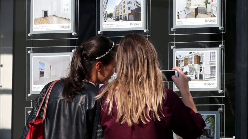 Two women view property listings in an estate agents window, one takes a photo with her phone. Real estate, stamp duty.