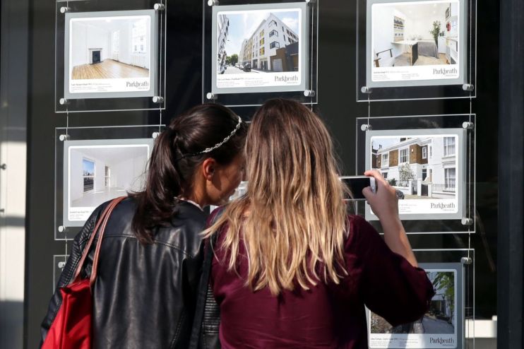 Two women view property listings in an estate agents window, one takes a photo with her phone. Real estate, stamp duty.