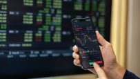 Hands holding a smartphone displaying a trading platform with cryptocurrency charts and buy/sell buttons, a blurred monito...