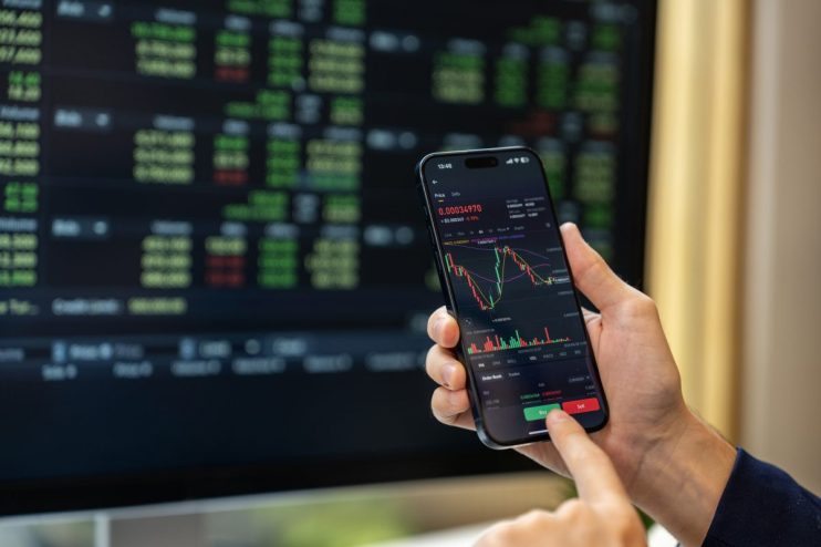 Hands holding a smartphone displaying a trading platform with cryptocurrency charts and buy/sell buttons, a blurred monito...