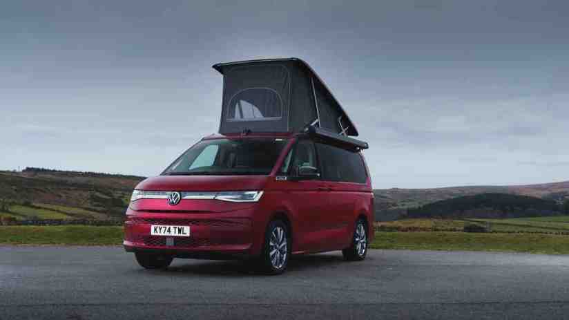 Red Volkswagen VW California camper van with pop-top roof extended, parked on a road with hills in background.