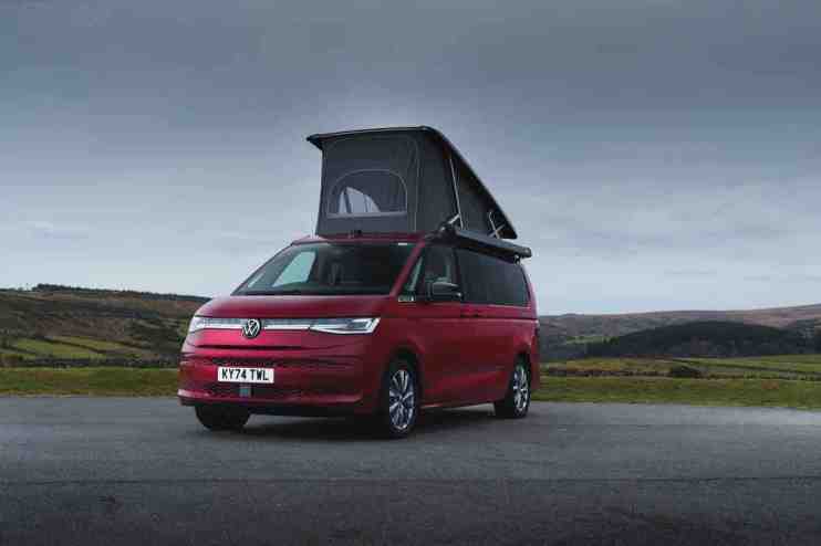 Red Volkswagen VW California camper van with pop-top roof extended, parked on a road with hills in background.