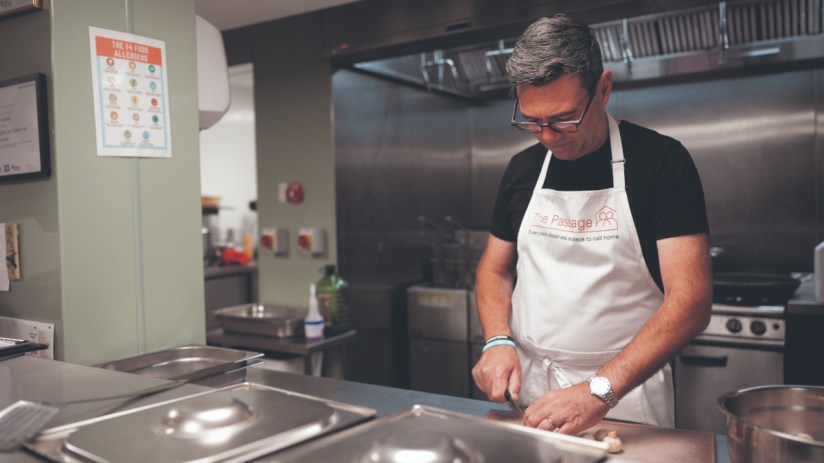 Man in The Passage apron slicing food in a commercial kitchen, with a food allergens chart visible.