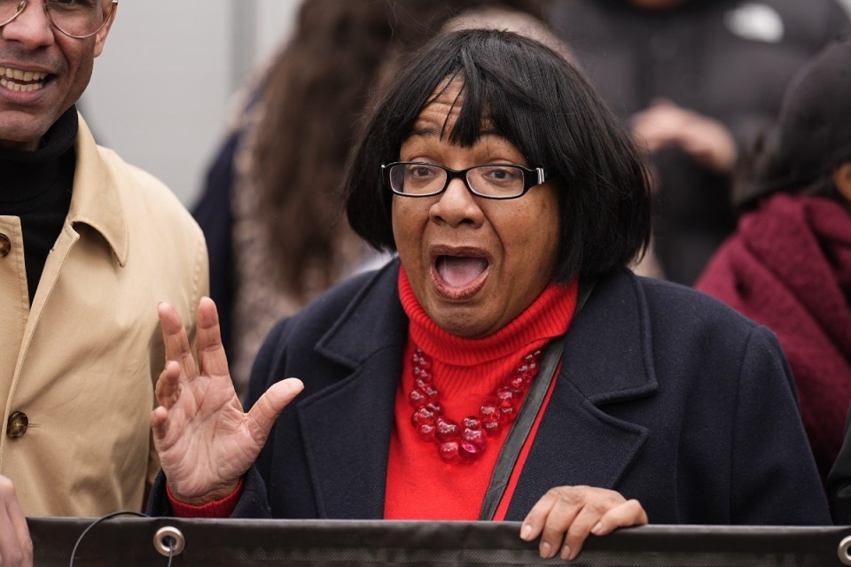 Woman in black coat and red turtleneck, glasses, speaking with wide-open mouth, next to a man in a tan coat.