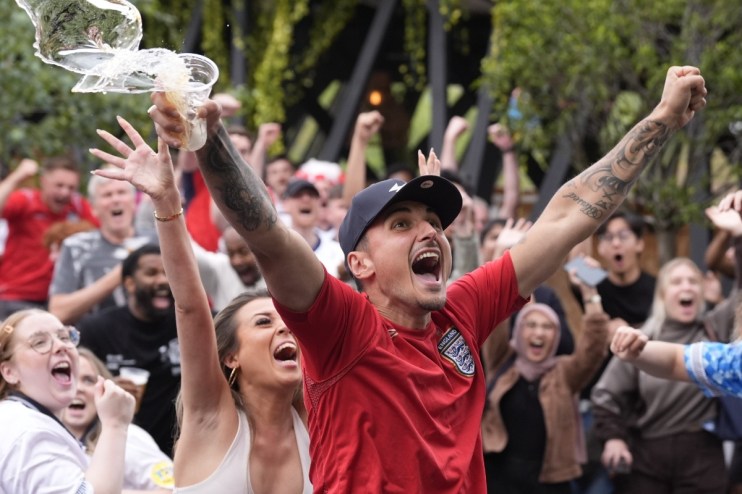 Excited crowd celebrating, a man in an England jersey cheers with arms raised and beer splashing from a cup.