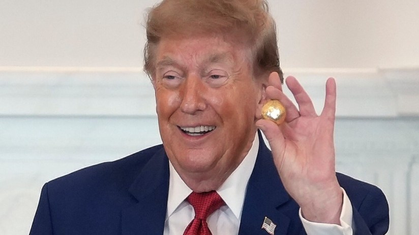 Donald Trump smiling and holding a small golden ball, wearing a blue suit and red tie.
