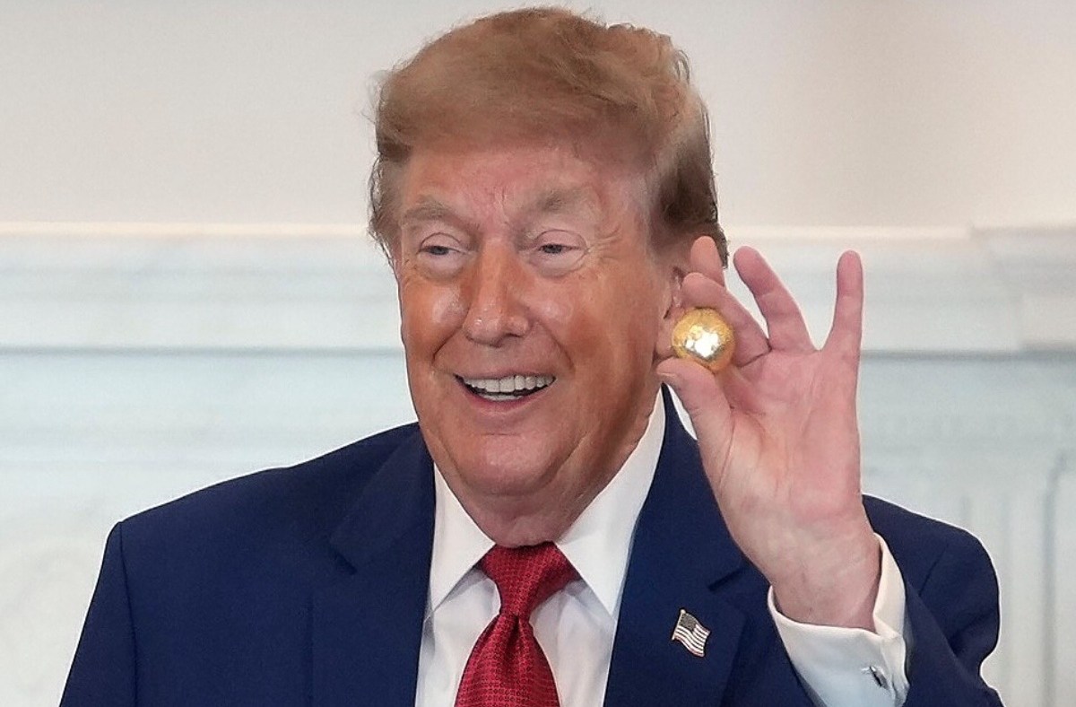 Donald Trump smiling and holding a small golden ball, wearing a blue suit and red tie.