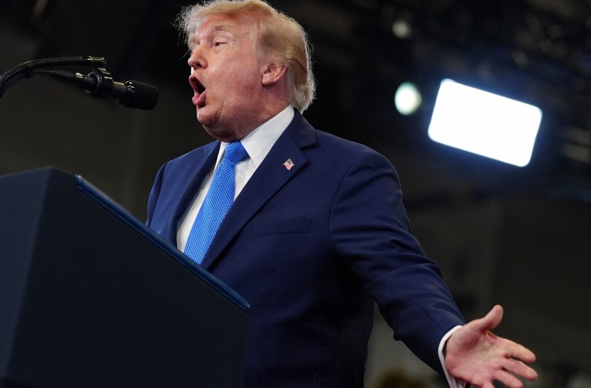 Donald Trump speaking emphatically at a podium, wearing a navy suit and blue tie, with a microphone and lights visible.