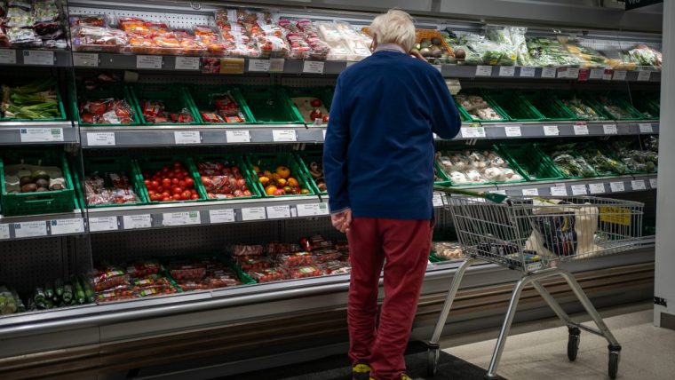 Shopper in a supermarket produce aisle browsing various packaged vegetables and fruits.