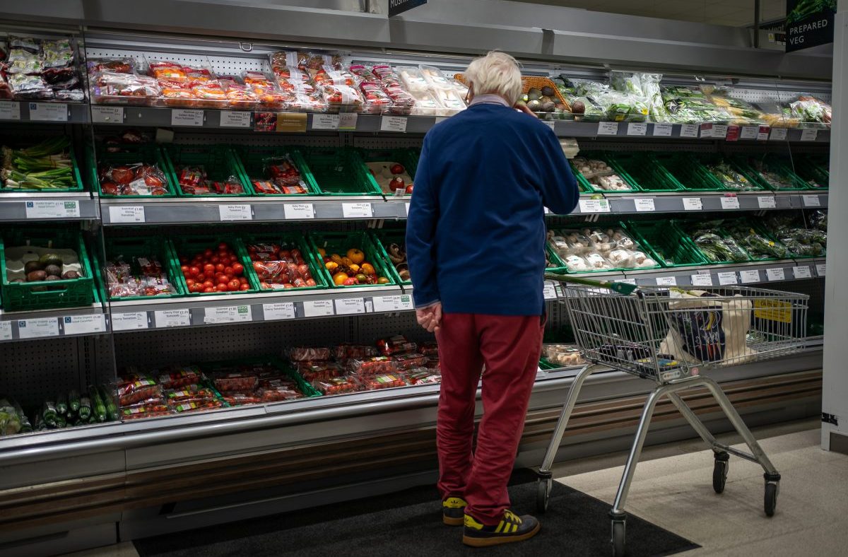 Shopper in a supermarket produce aisle browsing various packaged vegetables and fruits.