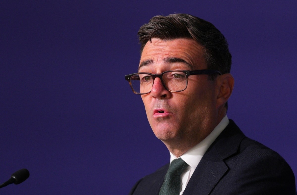 Andy Burnham, Mayor of Greater Manchester, speaking at a podium against a dark blue background.