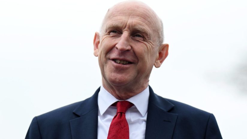 A smiling man in a dark suit and red tie looking slightly upwards, against a plain background.