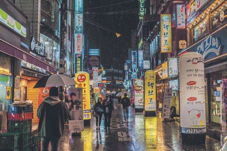 Rainy night street in Seoul, South Korea, with neon signs, storefronts, and people with umbrellas