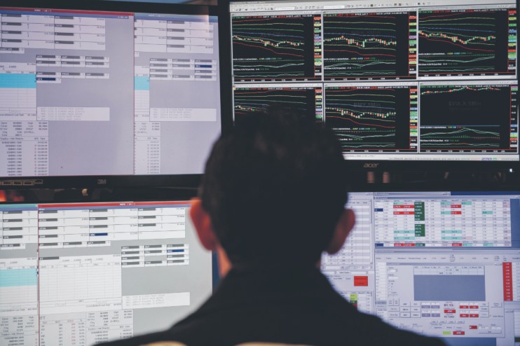 Trader monitoring multiple computer screens displaying stock market data, charts, and financial figures.