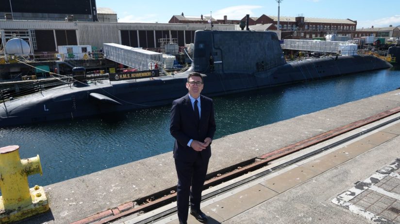 Andy Burnham in a suit, standing on a dock with the HMS Agamemnon submarine and BAE shipyard in the background.