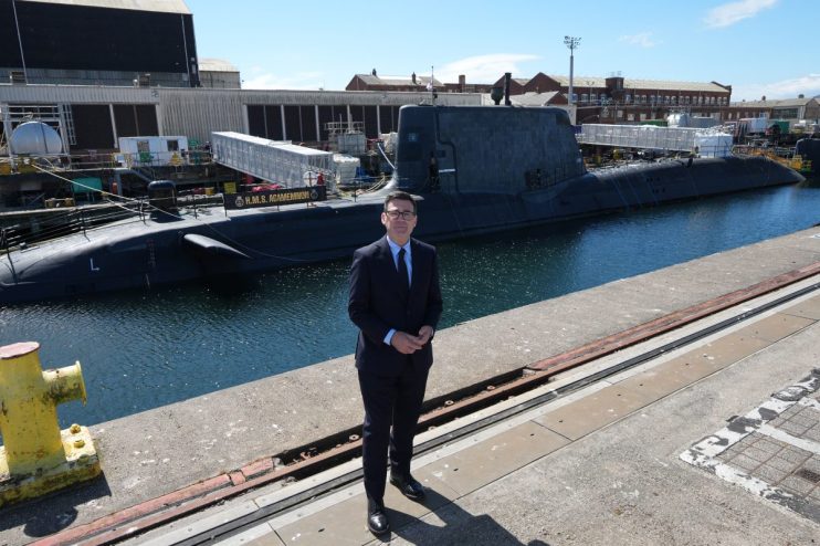 Andy Burnham in a suit, standing on a dock with the HMS Agamemnon submarine and BAE shipyard in the background.