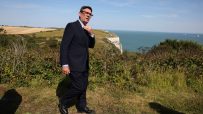 Andy Burnham adjusting his tie, overlooking white cliffs and the sea on a sunny day