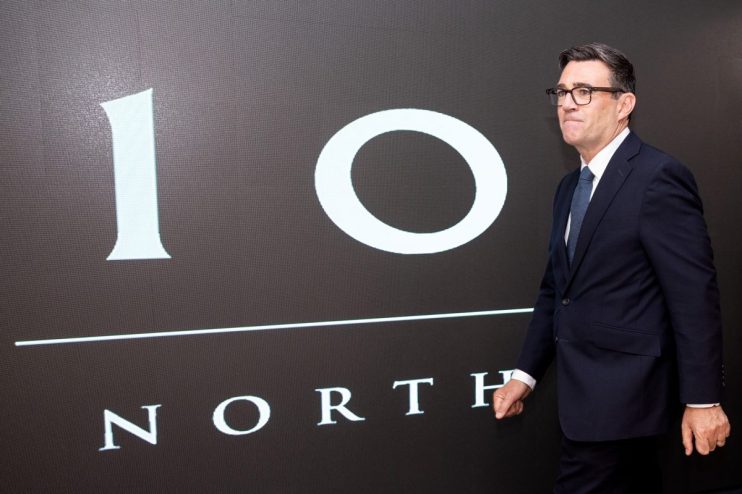 Andy Burnham in a dark suit and glasses, standing before a 10 NORTH graphic display.