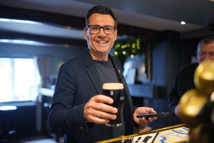 Andy Burnham smiling and holding a pint of beer and a smartphone in a pub setting
