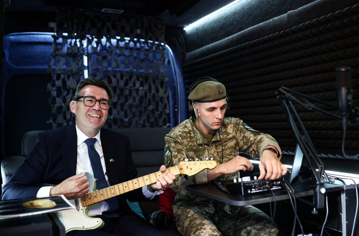 Andy Burnham smiling and playing guitar in Ukraine next to a soldier in uniform adjusting audio equipment