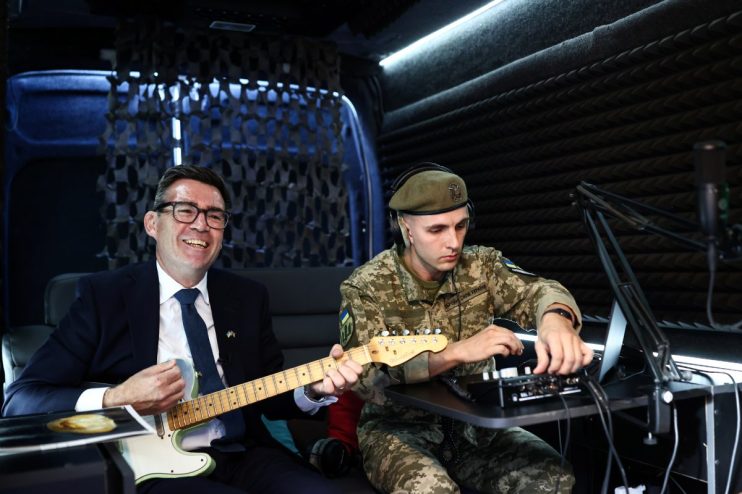 Andy Burnham smiling and playing guitar in Ukraine next to a soldier in uniform adjusting audio equipment