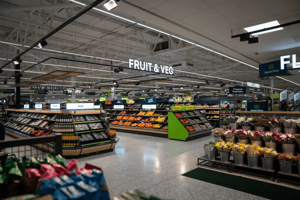 Asda supermarket aisle with Fruit & Veg sign, fresh produce displays, and flowers, reflecting Q2 trading