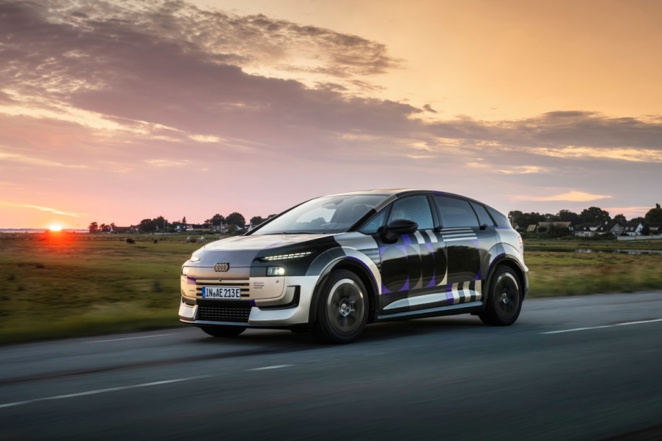 Audi A2 2 car with camouflage design driving on a road at sunset, fields and houses in background.