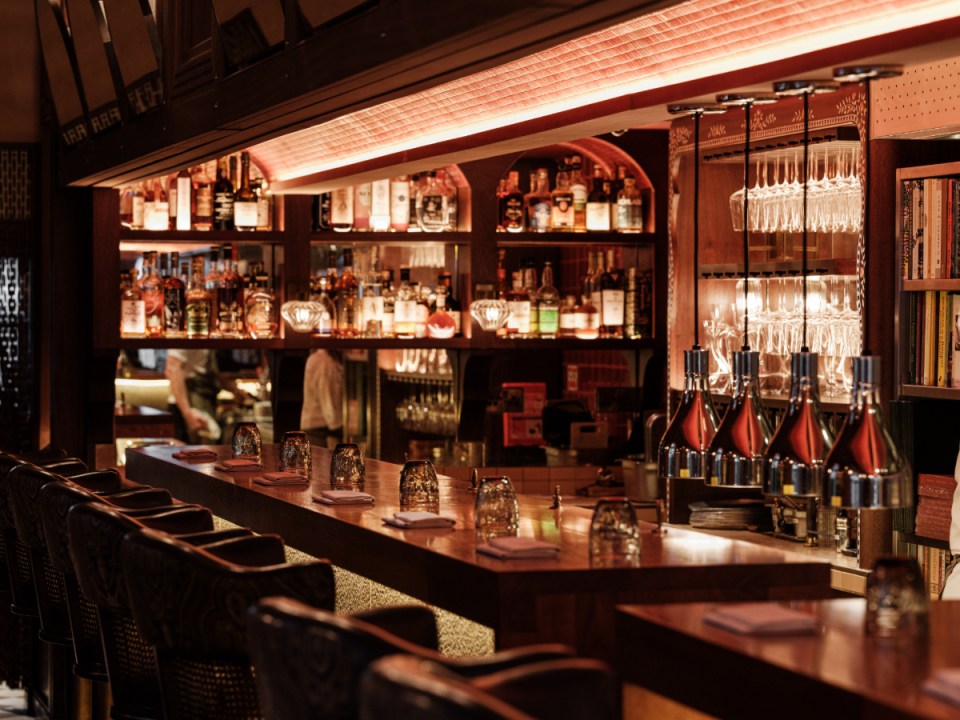 Warmly lit interior of BiBi restaurant bar with rows of bottles, glasses, and bar stools.