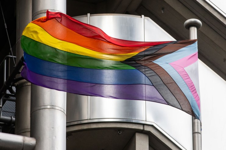 Progress Pride flag flying on a pole against a modern building, symbolizing trans policies in city firms