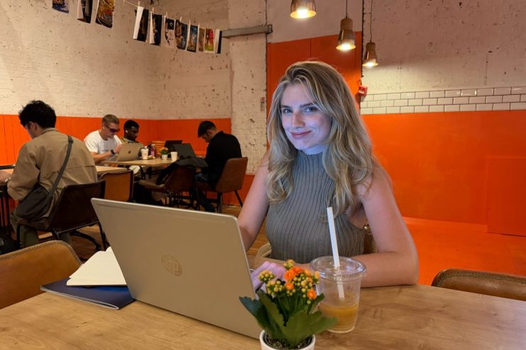 Anna, a young woman, smiles while typing on a laptop at a cafe table with a drink and potted plant.