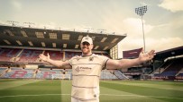 Smiling man in Burnley FC kit with Finotive One sponsor, arms outstretched, on a football pitch with stadium stands behind.