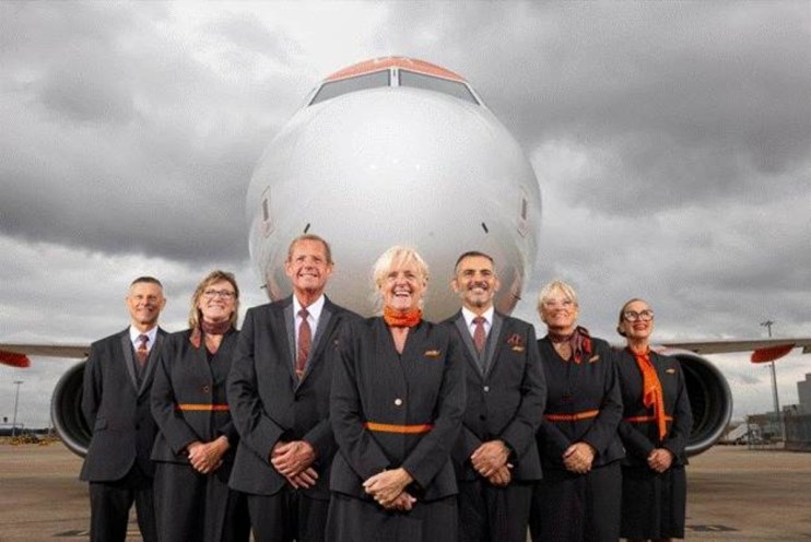 Six smiling EasyJet cabin crew, 50+, in uniform in front of an airplane on a cloudy day.