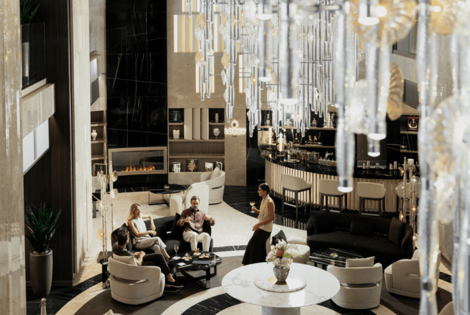 Elegant hotel lobby with guests relaxing, a bar, fireplace, and modern chandelier.