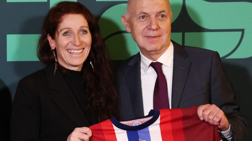 A smiling woman and a man in a suit holding a red, white, and blue soccer jersey with the Norway emblem.