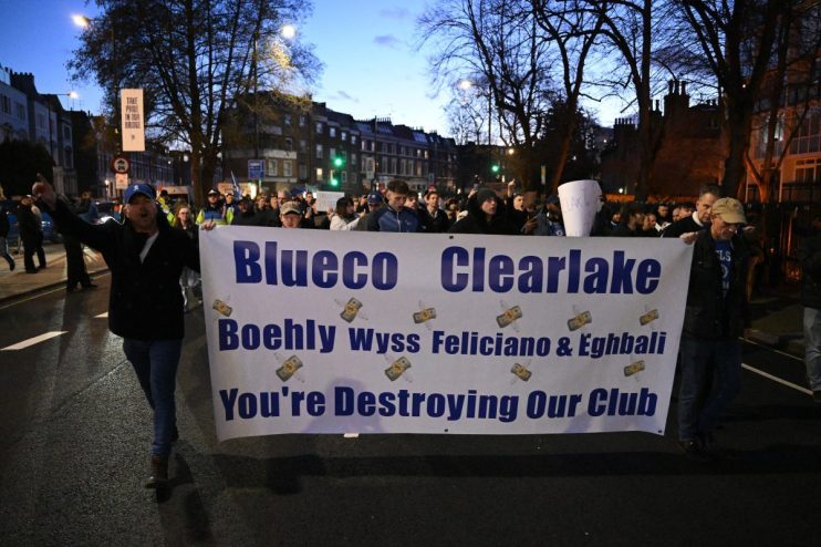 Football fans protest in the street holding a banner: Blueco Clearlake Boehly Wyss Feliciano & Eghbali Youre Destroying Ou...