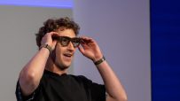 Mark Zuckerberg smiling while adjusting smart glasses on his face, wearing a black shirt and two wristbands