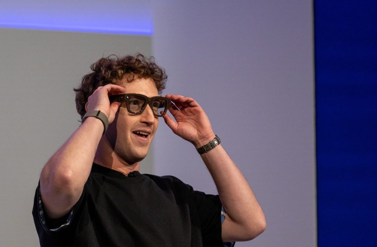 Mark Zuckerberg smiling while adjusting smart glasses on his face, wearing a black shirt and two wristbands