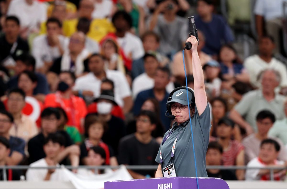 Female official in headset and hat raising a starting pistol at a sporting event, crowd blurred in background