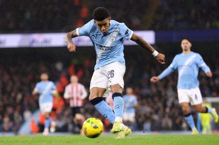 A male footballer in a light blue Manchester City kit, number 26, kicks a yellow soccer ball on a green field.