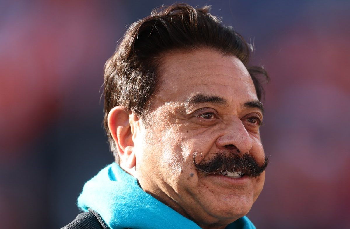 Shahid Khan, businessman, smiling and wearing a bright blue scarf, looking to the right with a blurred background.