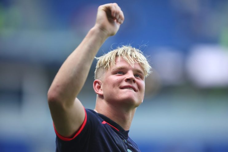 A young man with blonde hair and a dark shirt, clenching his fist and smiling, likely celebrating or cheering.