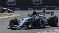 Blue Formula 1 race car on track with AWS branding on the barrier, another car in the background.