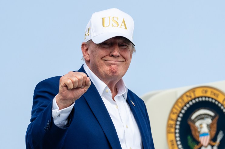 Donald Trump in a white USA hat, blue suit, and white shirt, making a fist, with a presidential seal visible