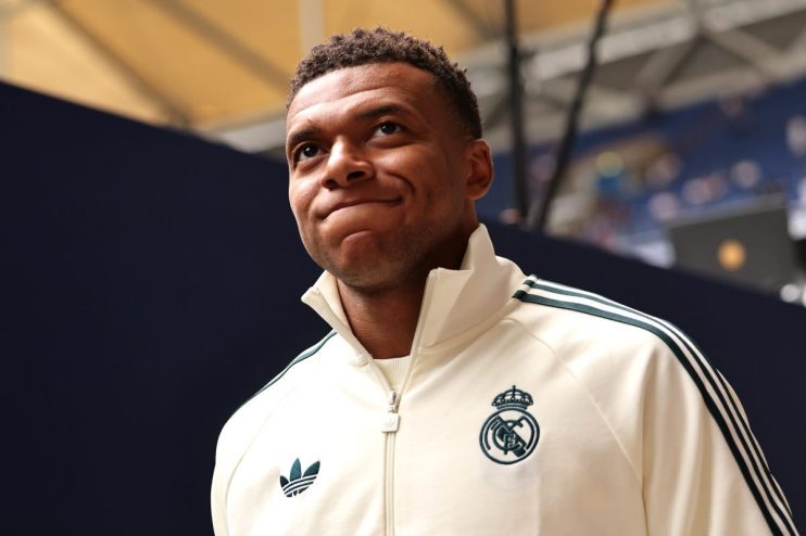 Kylian Mbapp&eacute; in a cream-colored Adidas track jacket with the Real Madrid crest, looking up with a thoughtful expression