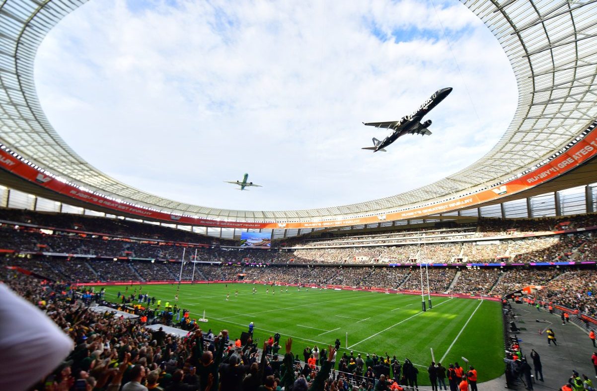 Two airplanes fly over a packed rugby stadium during a match, a large screen displaying Rugbys Greatest Rivalry