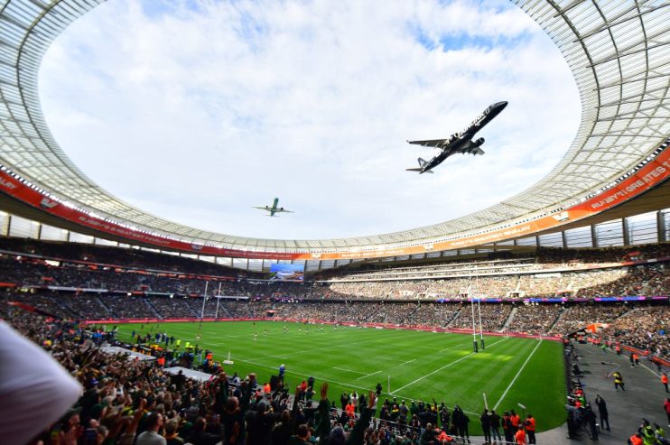 Two airplanes fly over a packed rugby stadium during a match, a large screen displaying Rugbys Greatest Rivalry