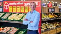 Giles Hurley, Aldi UK CEO, stands in a supermarket produce aisle with fresh fruits and vegetables.