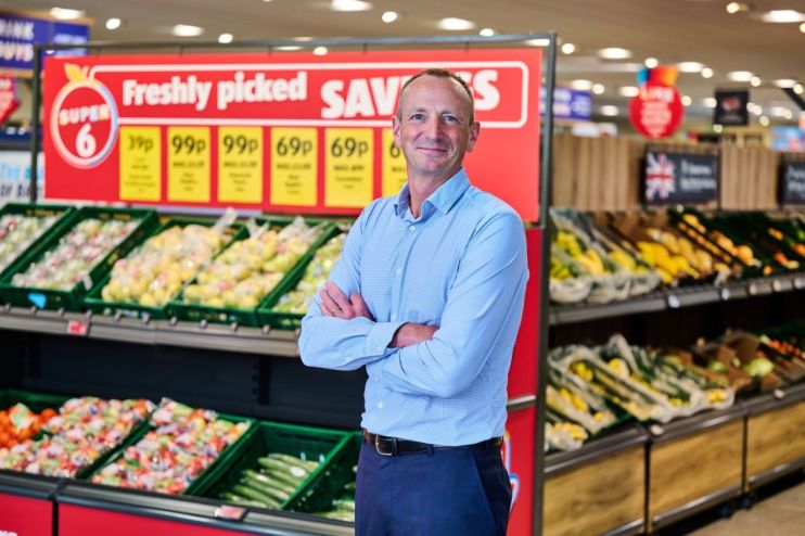 Giles Hurley, Aldi UK CEO, stands in a supermarket produce aisle with fresh fruits and vegetables.